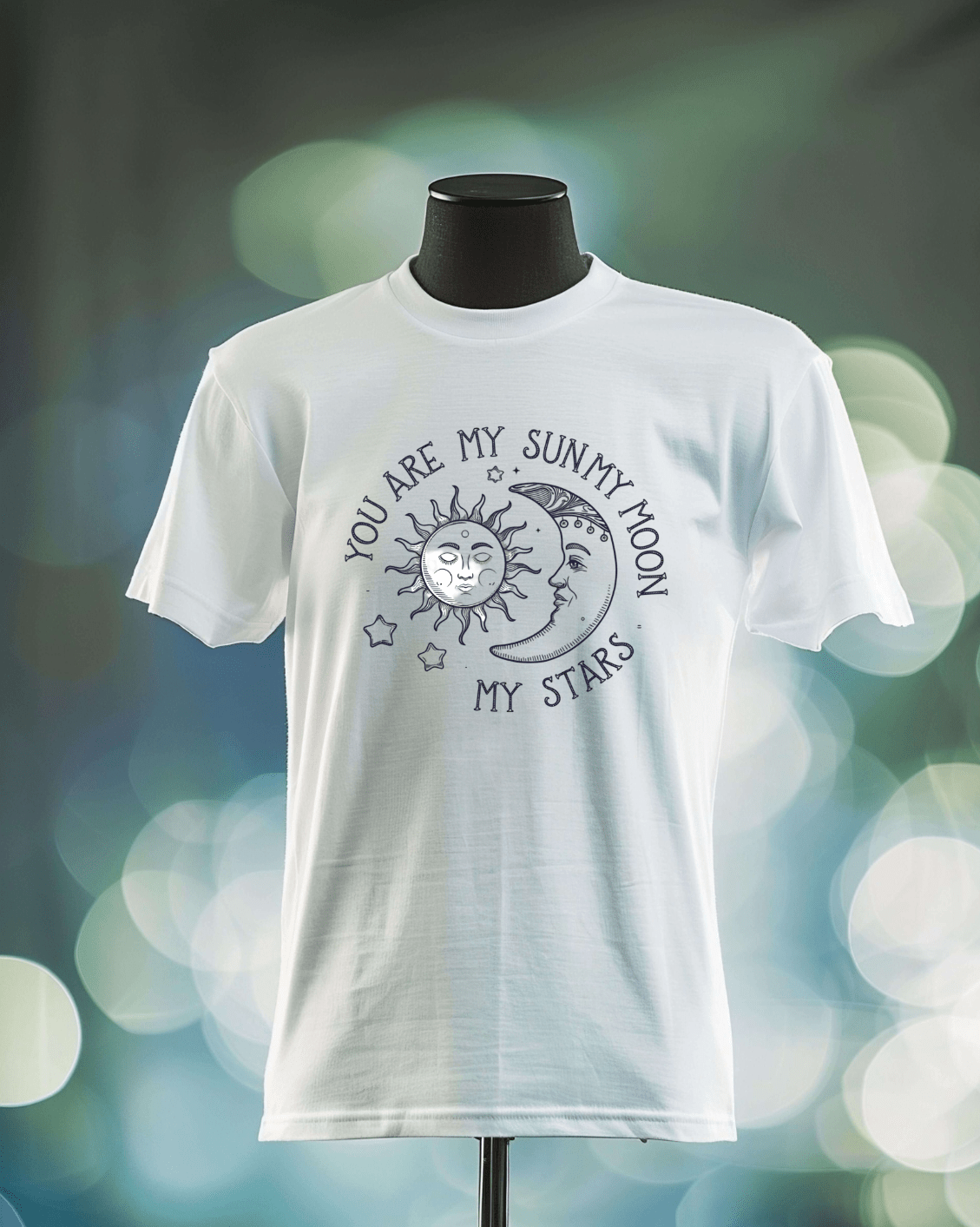 The Moon and Stars T-shirt by DB Custom Creations is shown on a mannequin with a sun and moon illustration and “You are my sun, my moon, my stars” in a circular pattern, printed in high-quality DTF. Soft bokeh highlights the display.