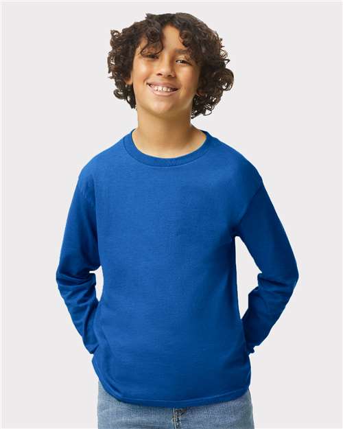 Smiling child with curly hair wears a bright blue Gildan 5400B long-sleeve shirt and light blue jeans, standing with hands in pockets against a plain light background.
