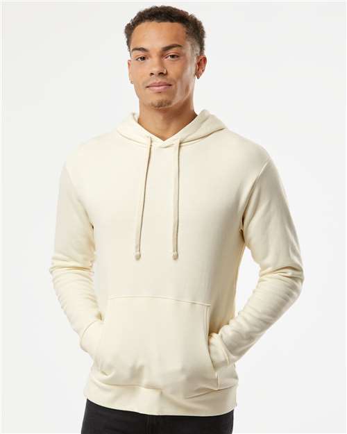A young man stands with his hands in the pockets of a cream-colored Next Level 9303 cotton polyester hoodie, featuring a jersey lined hood and front pocket, against a plain white background.