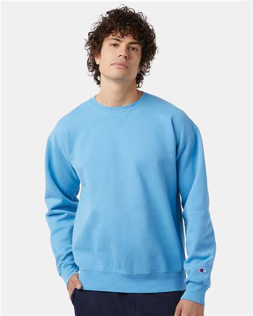 A person with curly hair wears a light blue Champion CD400 no-shrink cotton sweatshirt and dark pants, standing against a plain white background.