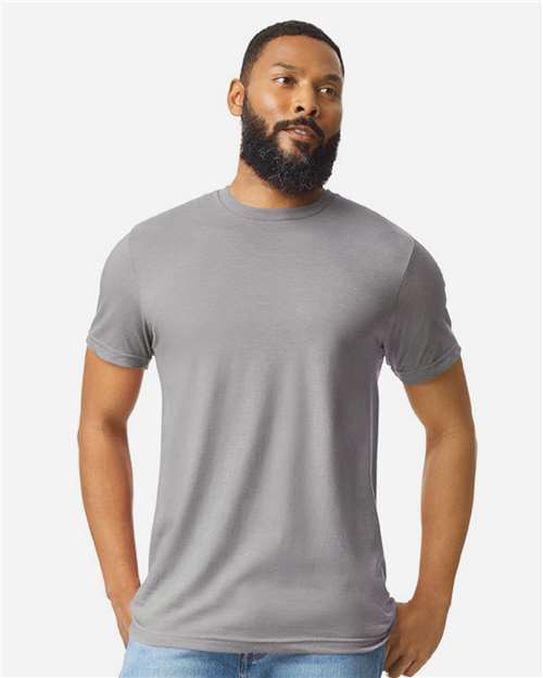 A bearded man with short hair is wearing a plain gray Gildan Softstyle 64000CVC t-shirt and light blue jeans, standing against a plain white background with his hands in his pockets.