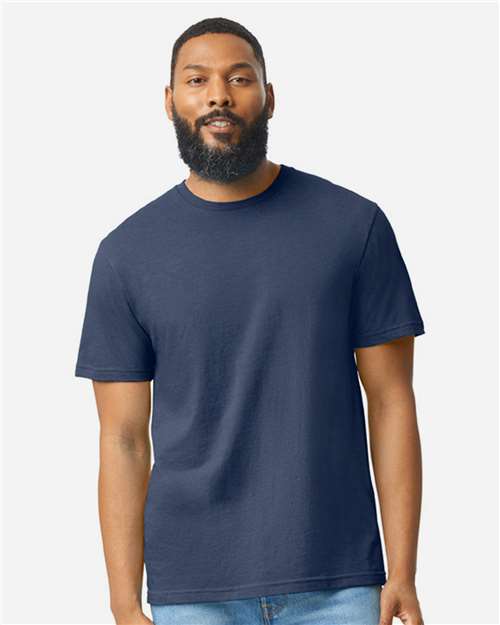 A man with a beard and short hair is wearing a plain navy blue Gildan Softstyle 64000CVC t-shirt by Gildan, made from ring-spun cotton, and light blue jeans, standing against a light gray background.