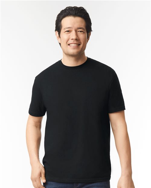 A dark-haired man smiles against a white background, wearing a plain black Gildan Softstyle 64000CVC t-shirt by Gildan and blue jeans.