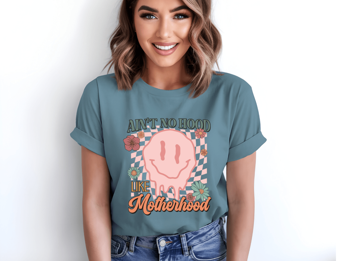 A smiling woman with shoulder-length hair wears the Aint no Hood like Motherhood T-shirt by DB Custom Creations, featuring a vibrant pink melting smiley face, flowers, checkered pattern, and bold text using vivid DTF printing.