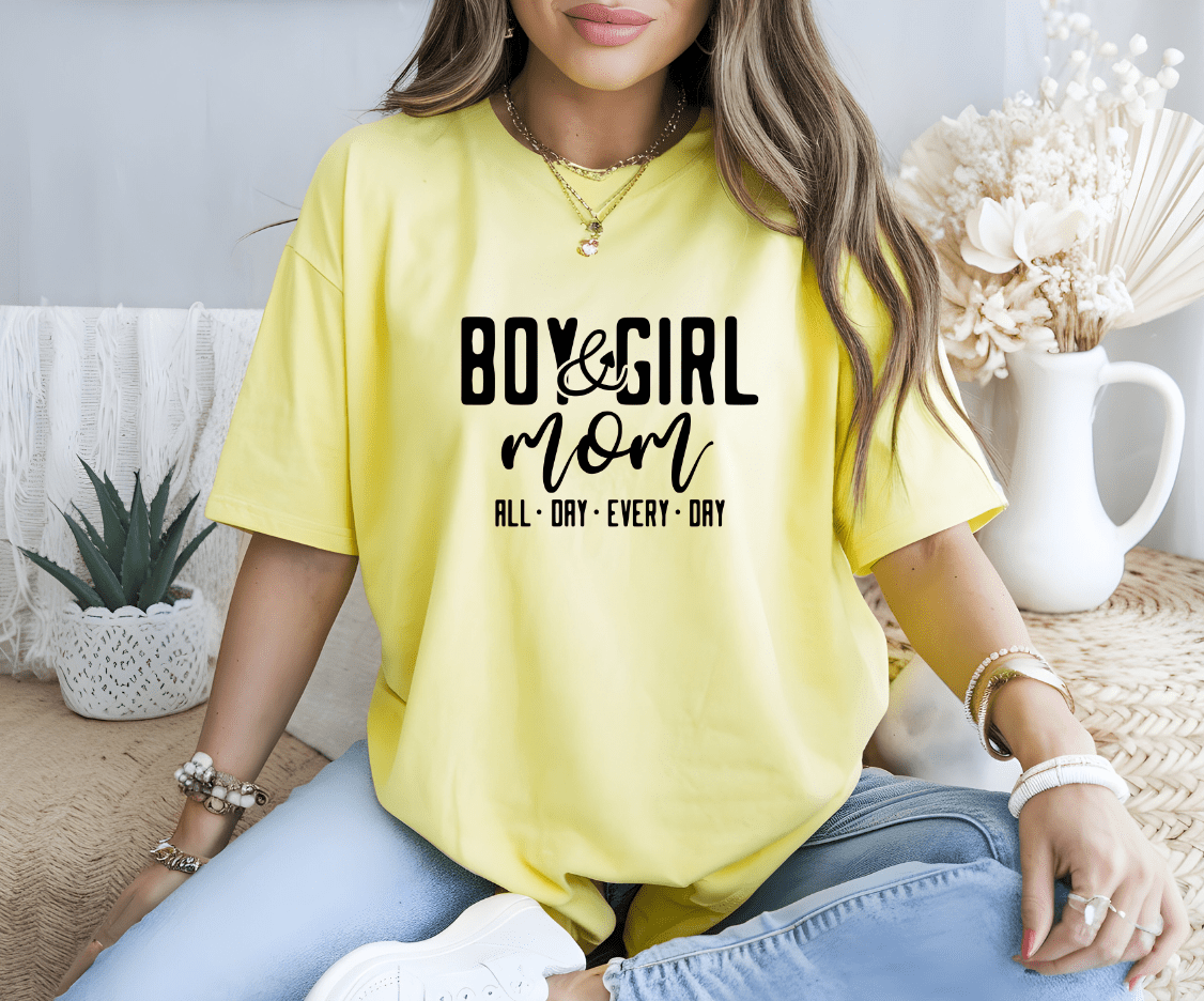 A woman wears a yellow Boy and Girl Mom DTF Transfer shirt by DB Custom Creations, sitting cross-legged on the floor with bracelets and a necklace. Dried flowers and a potted plant decorate the background.