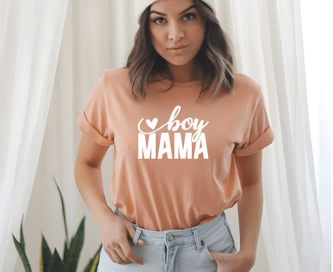 A woman indoors, in soft light with white curtains and a plant, wears the peach-colored Boy mama T-shirt by DB Custom Creations, featuring boy MAMA and a heart design created with 11-inch DTF printing.