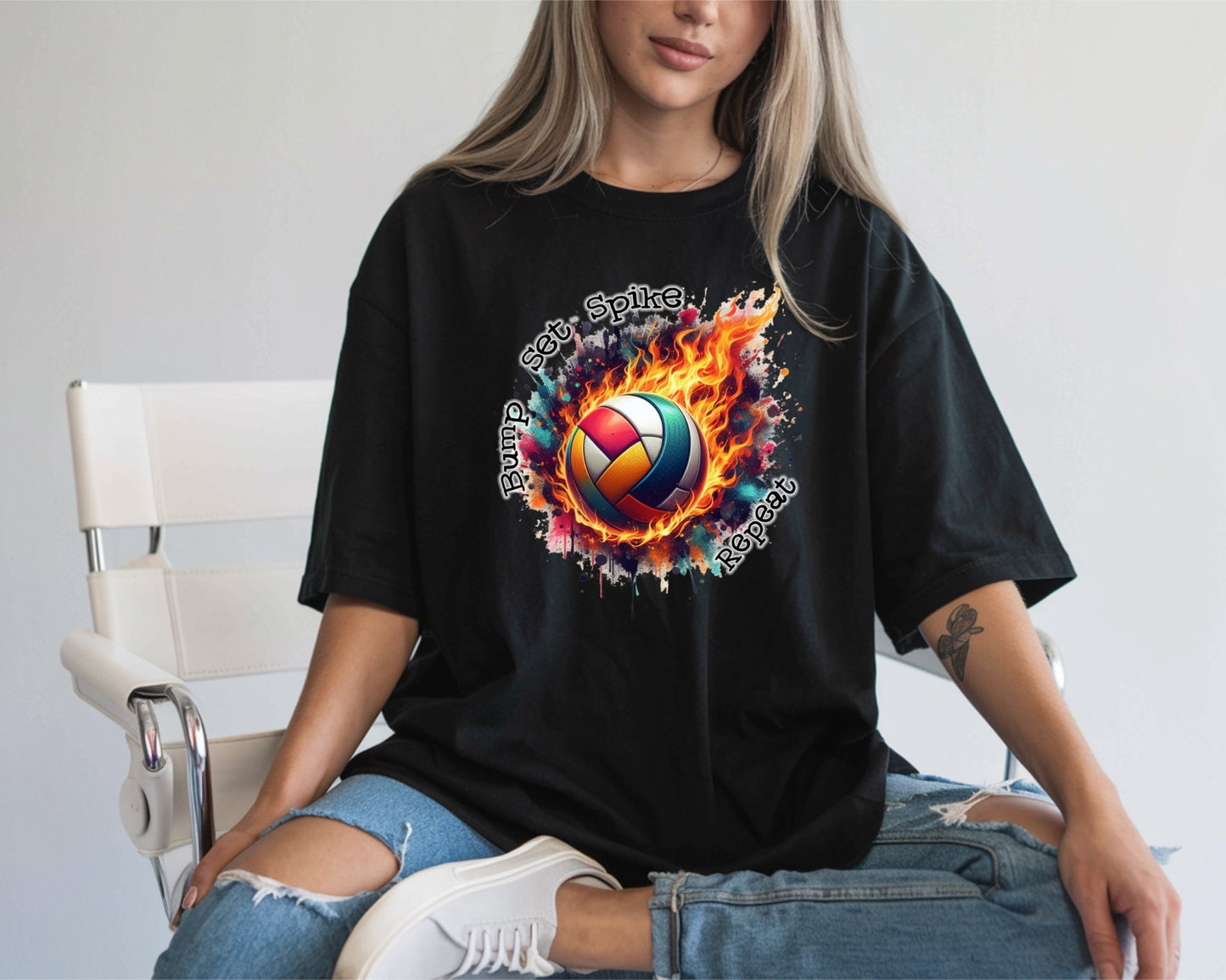 A woman with long blonde hair and ripped blue jeans sits on a white chair wearing the DB Custom Creations “Bump Set Spike” black t-shirt, made with DTF printing and featuring a flaming volleyball graphic.