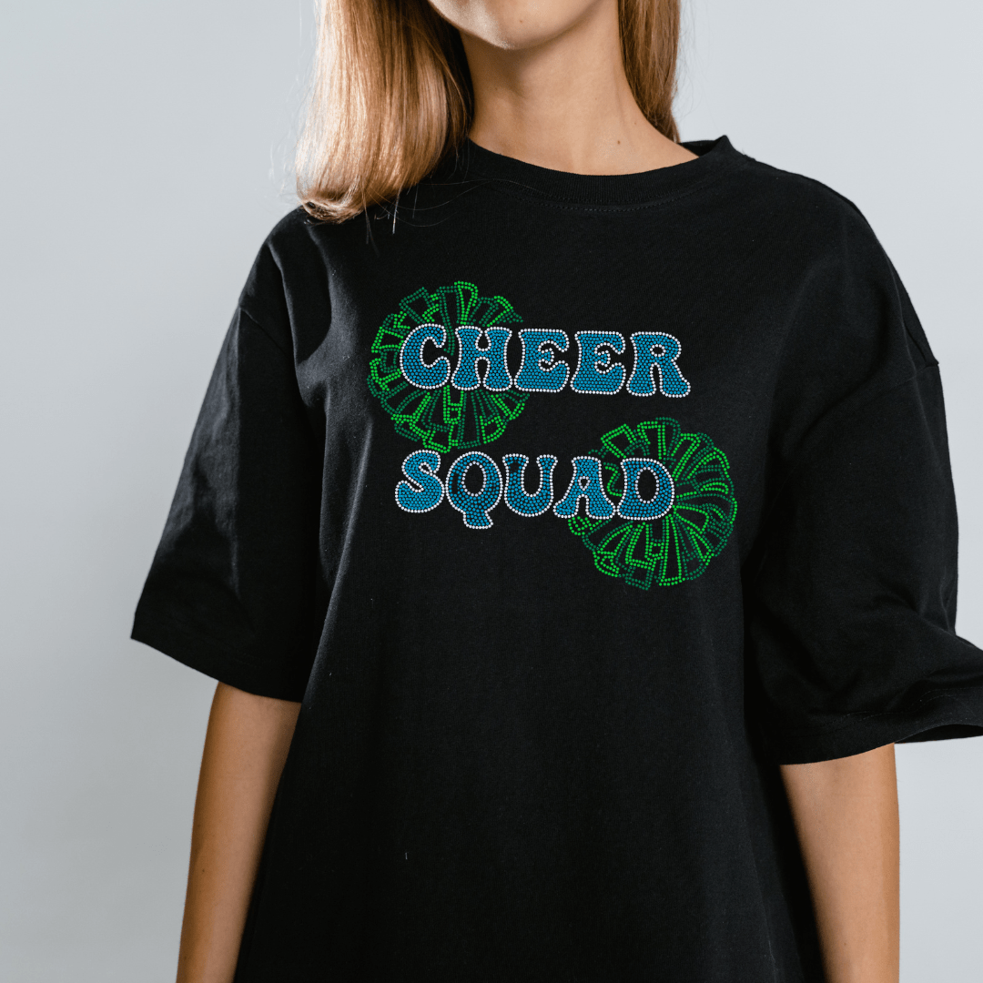 A woman wears a DB Custom Creations Spangle Transfer - Teal Cheer Squad black t-shirt with blue and white lettering and green pom-pom graphics. Her head is partially out of frame against a plain, light background.