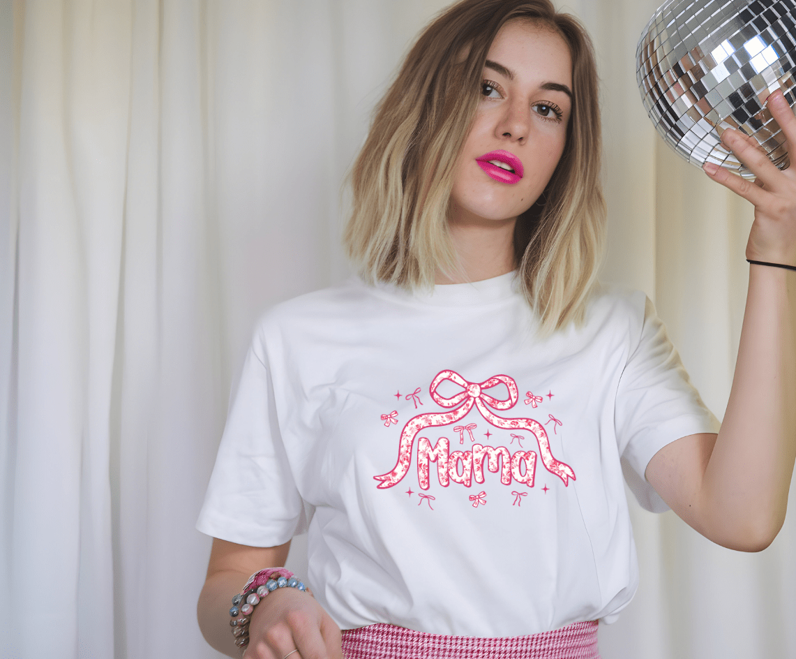 A young woman with shoulder-length blonde hair wears the Coquette Mama white T-shirt by DB Custom Creations, featuring a vibrant Mama and pink bow graphic. She pairs it with a pink skirt and colorful bracelets, holding a disco ball.