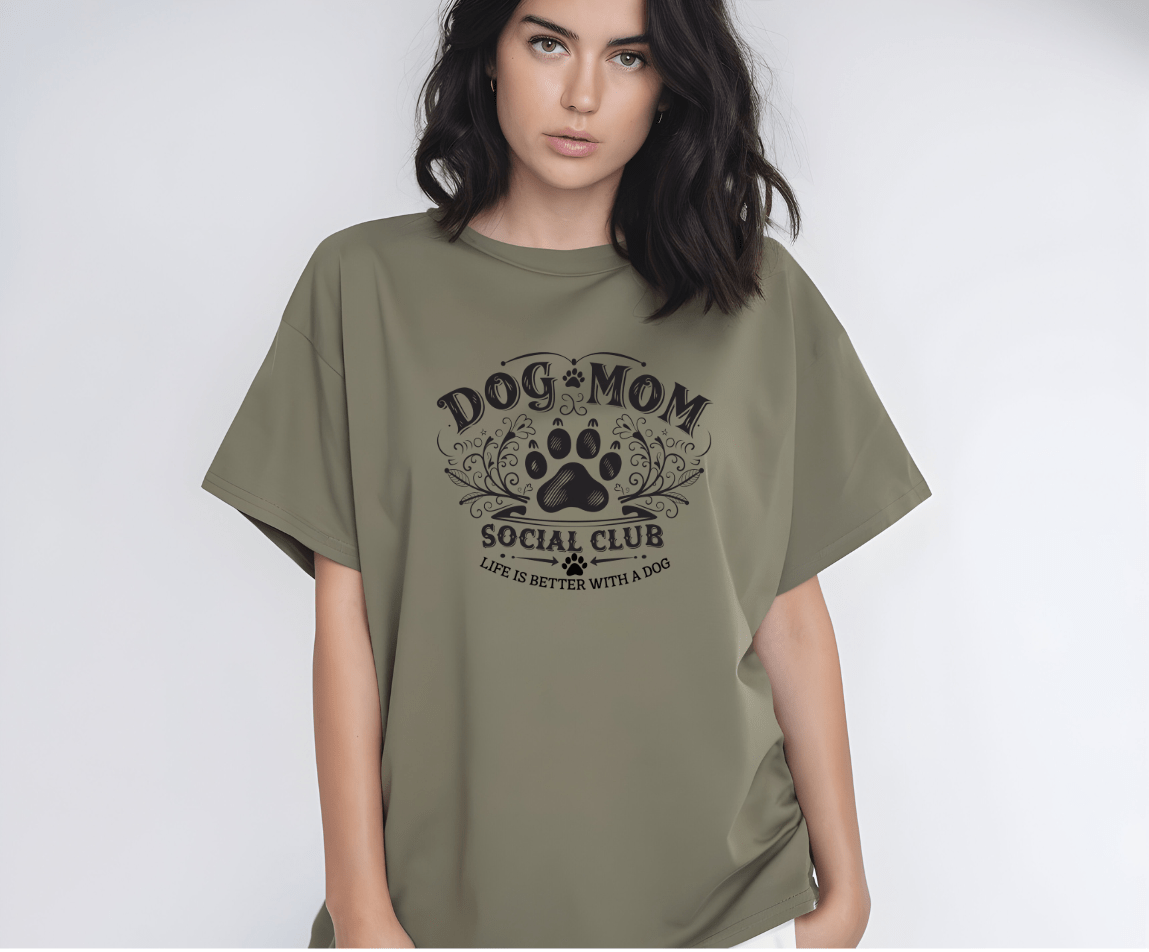 A woman with shoulder-length dark hair wears the Dog Mom olive-green t-shirt by DB Custom Creations, featuring DTF printing, a paw print graphic, and the slogan Dog Mom Social Club - Life is better with a dog, standing against a plain background.