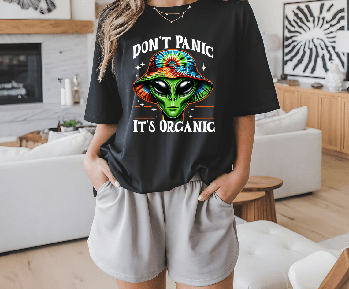 A person stands indoors in light gray shorts and a black Dont panic its Organic t-shirt by DB Custom Creations, featuring a vibrant DTF Transfer print of a colorful alien face with a tie-dye hat.