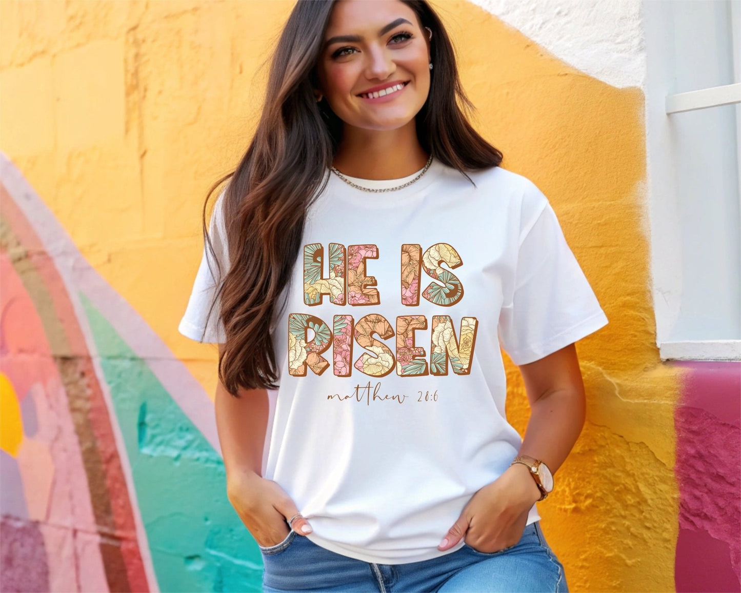 A smiling woman with long brown hair wears the “He Is Risen” white T-shirt by DB Custom Creations, featuring colorful DTF printing and the text “Matthew 28:6,” as she stands before a vibrant multicolored wall.