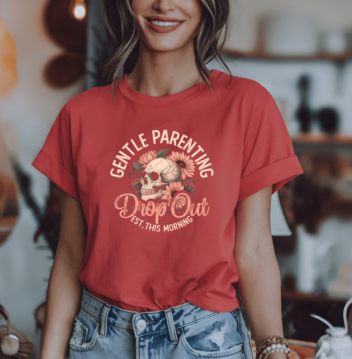 A woman indoors, smiling, wears a red rolled-sleeve t-shirt from DB Custom Creations featuring the Gentle Parenting Dropout design with a floral skull graphic.