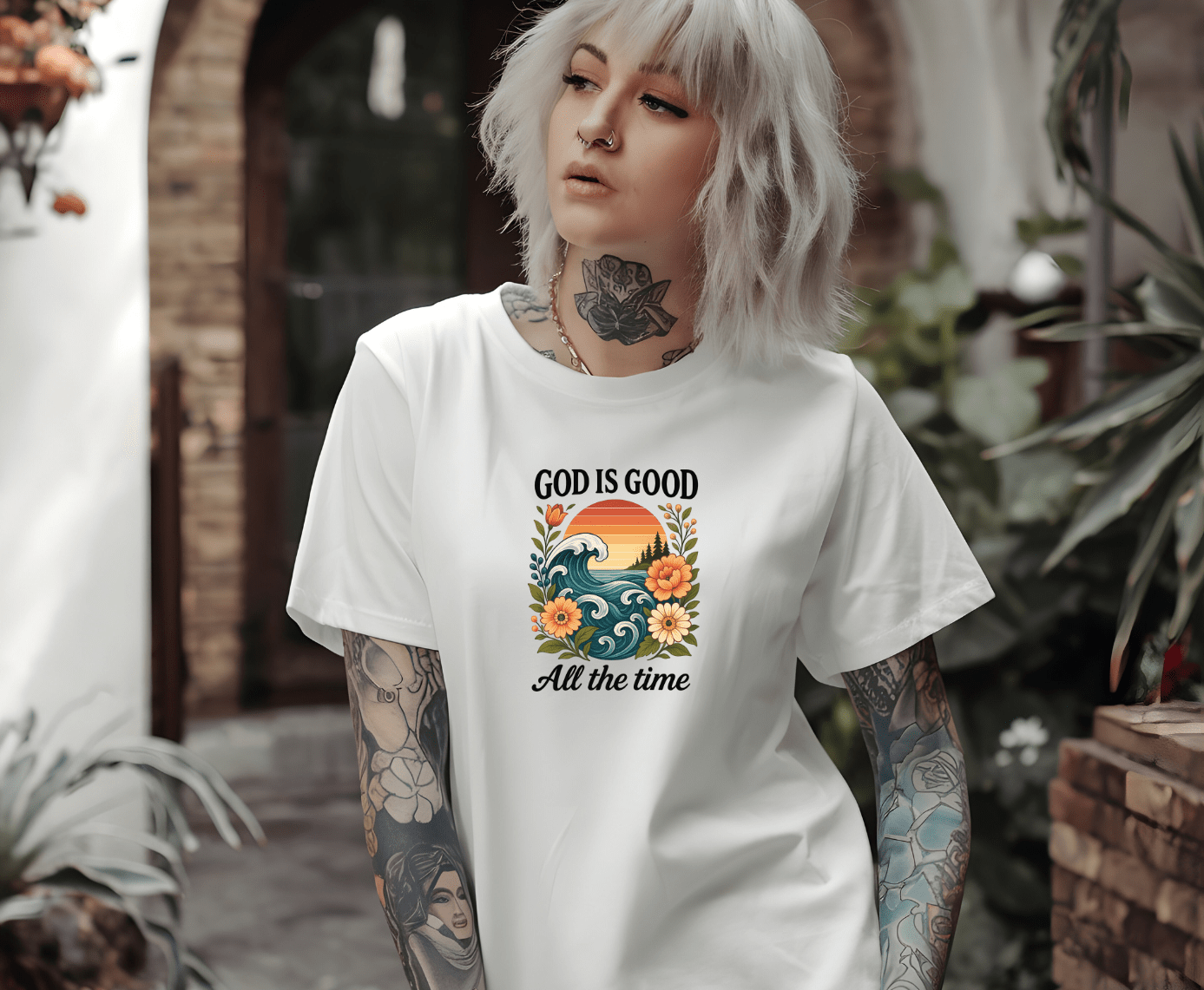 A gray-haired, tattooed woman wears a white T-shirt from DB Custom Creations featuring the God is good DTF Transfer design with waves, flowers, and a sunset. She stands outside near plants and brick walls.