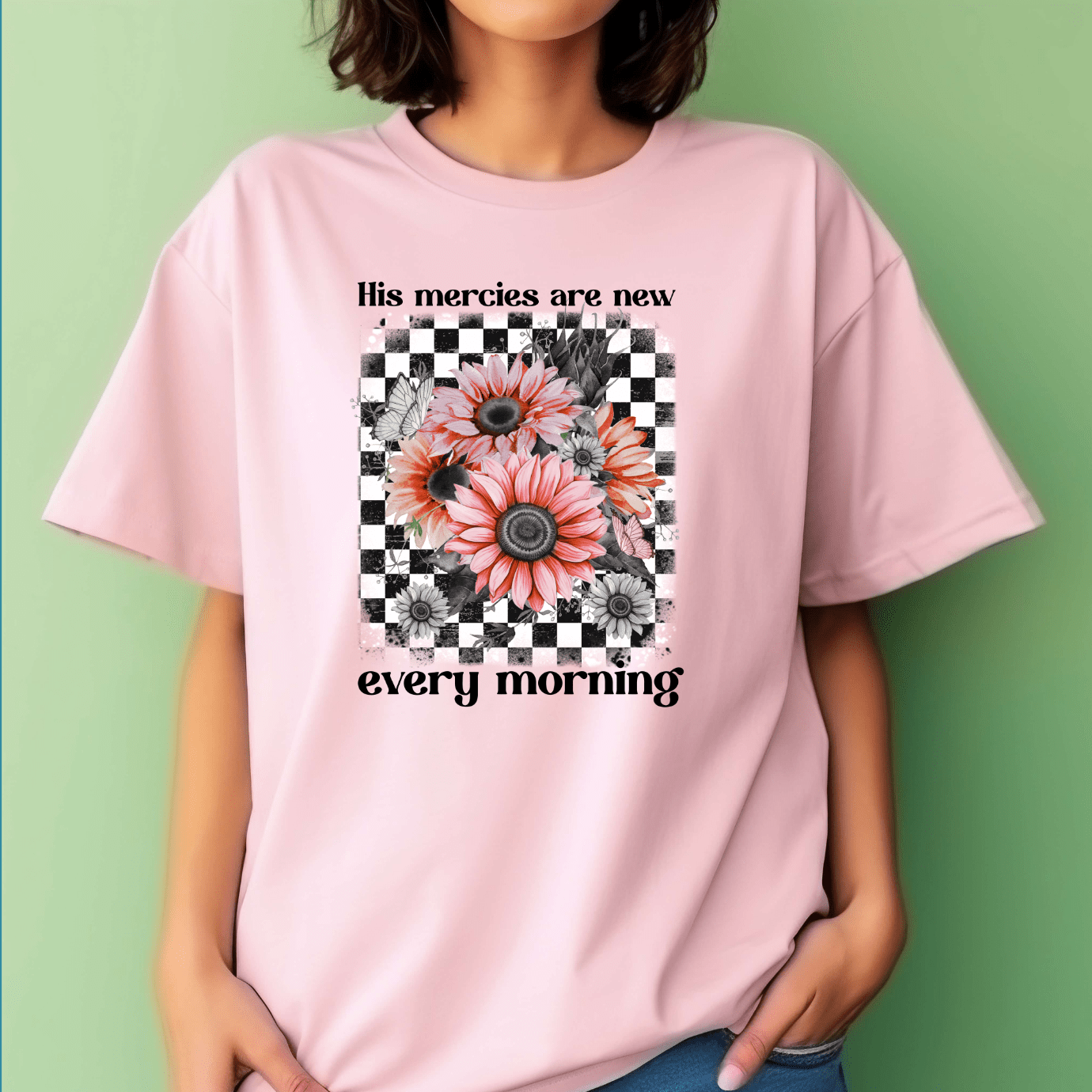 A woman wears the “His Mercies Are New” T-shirt by DB Custom Creations, featuring a checkered black-and-white square, pink sunflowers, and uplifting text on a pale pink shirt with an 11-inch DTF transfer. The light green background adds freshness.