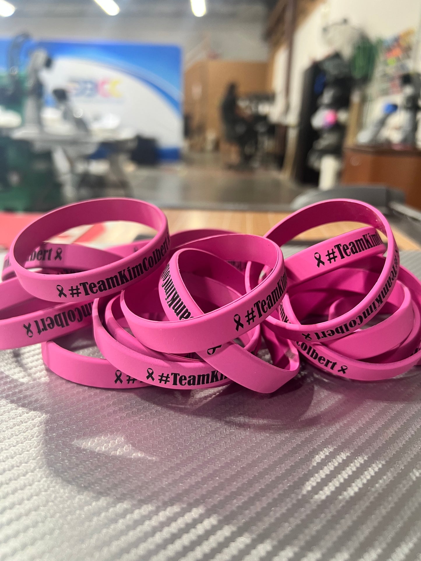 Custom Silicone Bracelets by DB Custom Creations featuring #TeamKimColbert and a pink ribbon, ideal for fundraising, are shown in a bright indoor setting on a textured surface.