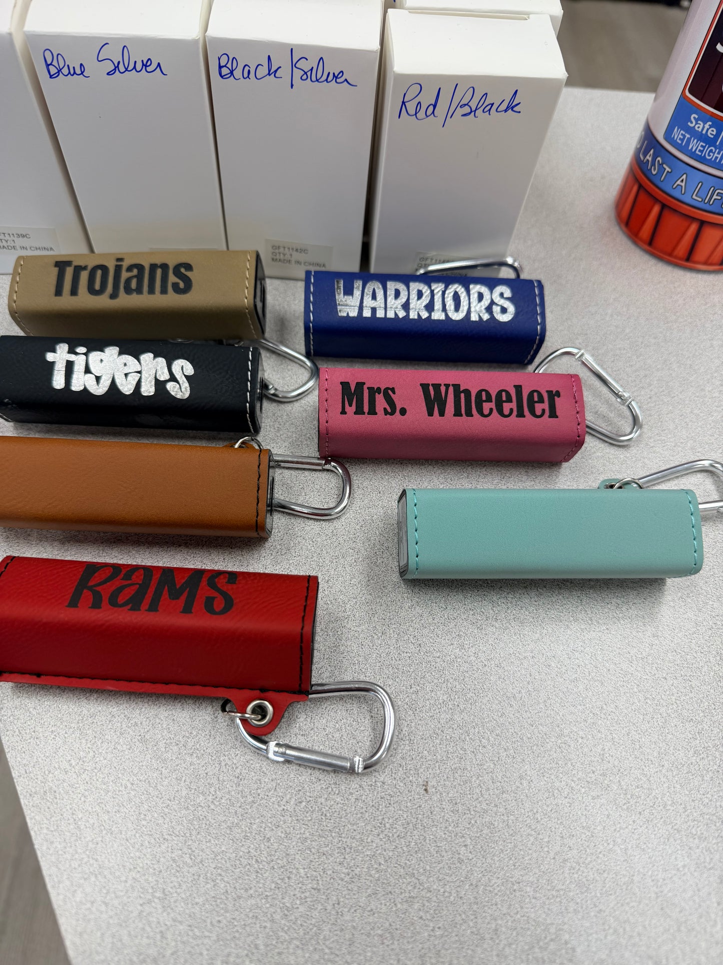 Six vibrant, rectangular Custom Laser Engraved Battery Banks by DB Custom Creations—each featuring different personalized names such as Trojans, Warriors, Tigers, Mrs. Wheeler, RAMS, and a mint green design—are displayed on a table alongside labeled boxes in the background, making them an ideal personalized giveaway or accessory for any event.