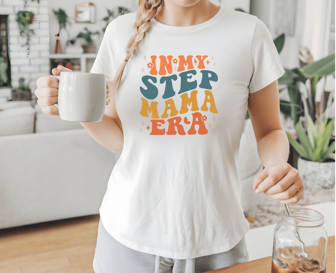 A woman wears a white Step mama era t-shirt by DB Custom Creations, holding a white mug in a cozy living room with plants and decor in the background.