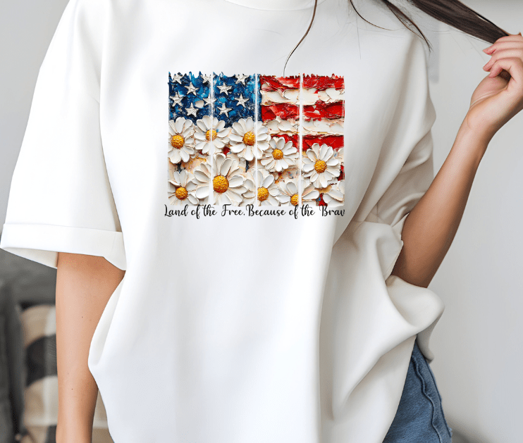 Someone wears the Land of the Free white shirt by DB Custom Creations, featuring a U.S. flag with daisy stripes and Land of the Free Because of the Brave text, made with vibrant DTF heat transfer.