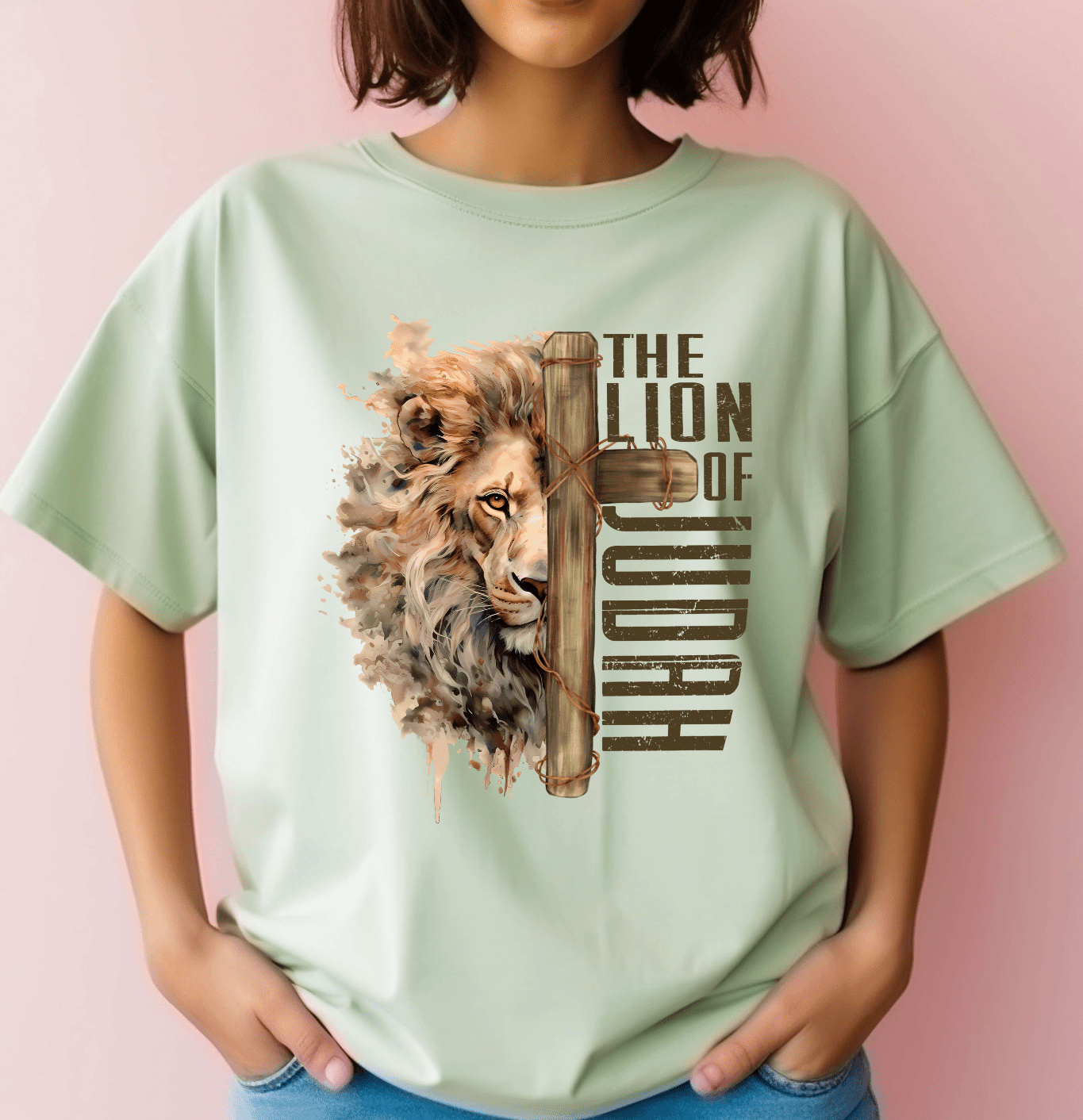 A person wears the Lion of Judah T-shirt by DB Custom Creations, featuring an illustrated lion’s face, a scroll, and THE LION OF JUDAH text in DTF Transfer. They stand against a pale pink background, hands in jean pockets.