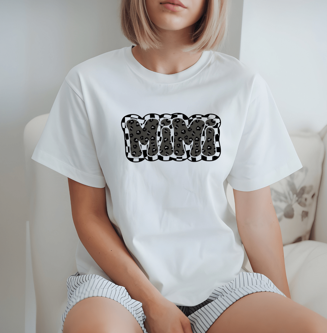 A woman with a bob haircut sits on a chair wearing the “Mimi” white t-shirt by DB Custom Creations, featuring bold bubble letters in a black and white spotted pattern made with DTF Printing, paired with striped shorts.