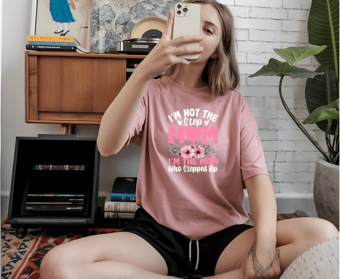 A woman sits on the floor taking a selfie, wearing the Mom that stepped up pink t-shirt by DB Custom Creations, featuring the phrase I’m not the step mom. I’m the mom who stepped up with floral and heart accents. A plant and wooden furniture are in the background.