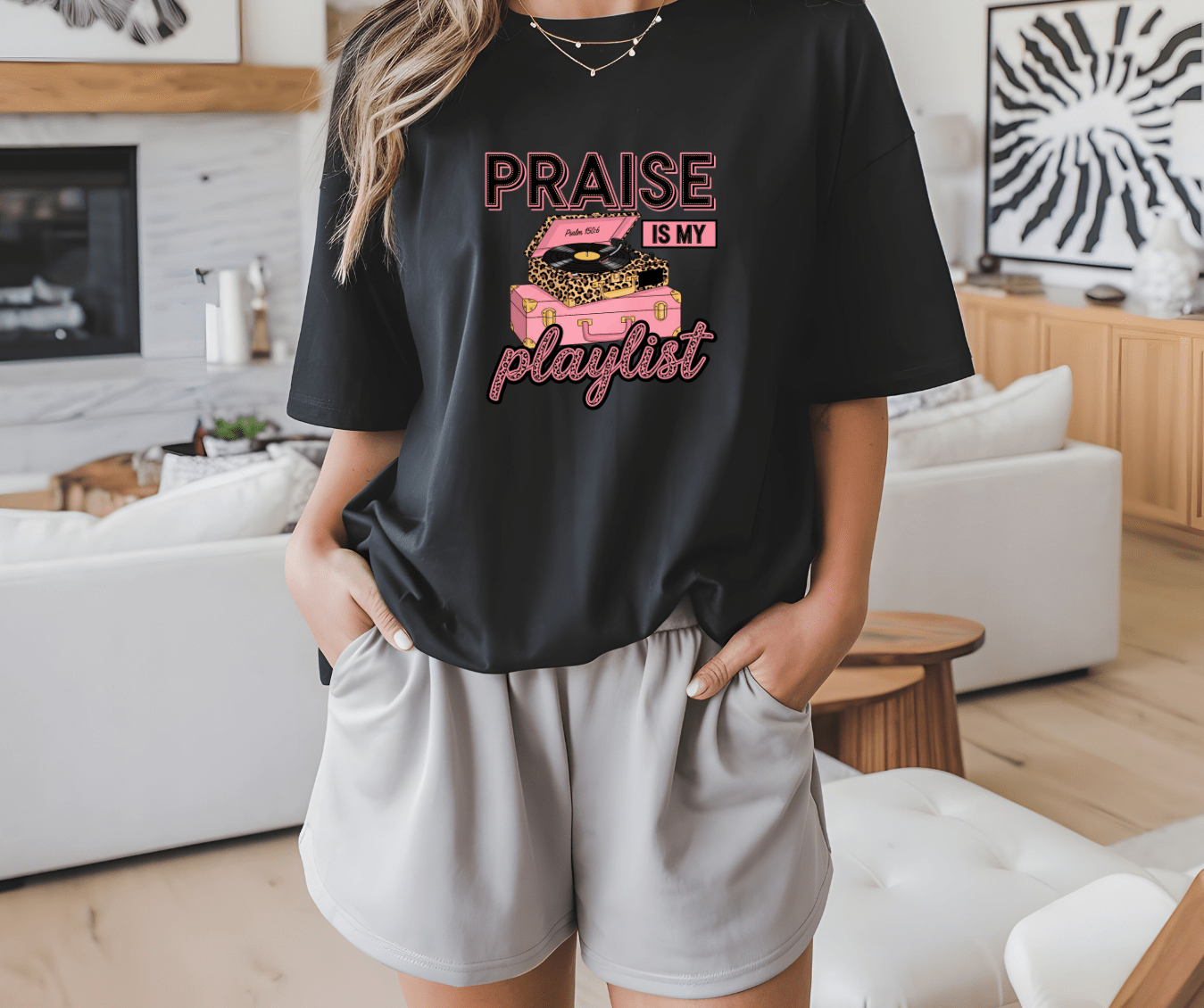 A person in a living room wears the DB Custom Creations Praise is my playlist black t-shirt with an illustrated pink record player, paired with light gray shorts, and has their hands in their pockets.
