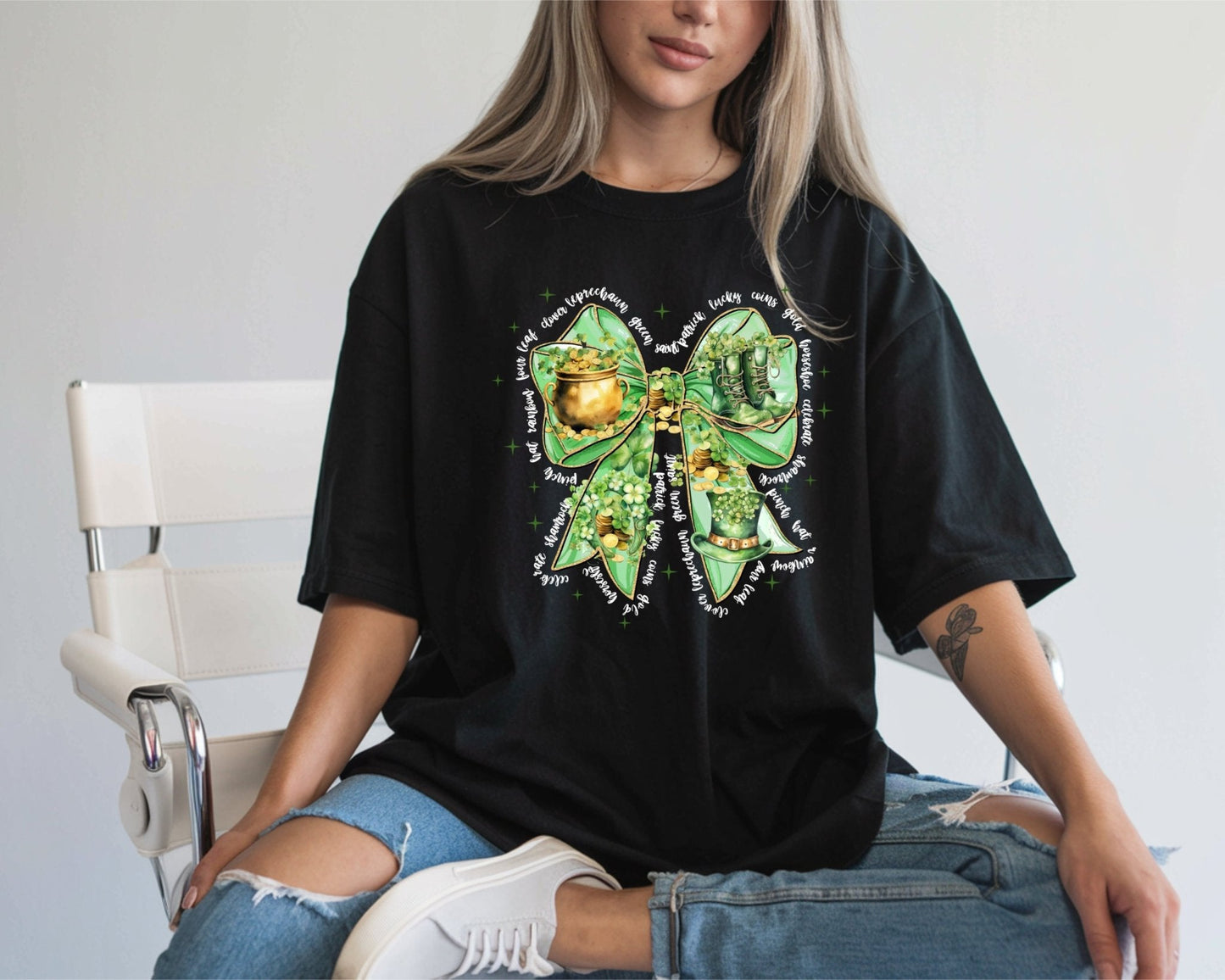 A woman sits on a white chair wearing a DB Custom Creations Bow with White Wording 11-inch DTF Transfer black t-shirt, featuring a large green bow with gold coins, shamrocks, and shoes. She pairs it with ripped blue jeans and white sneakers.