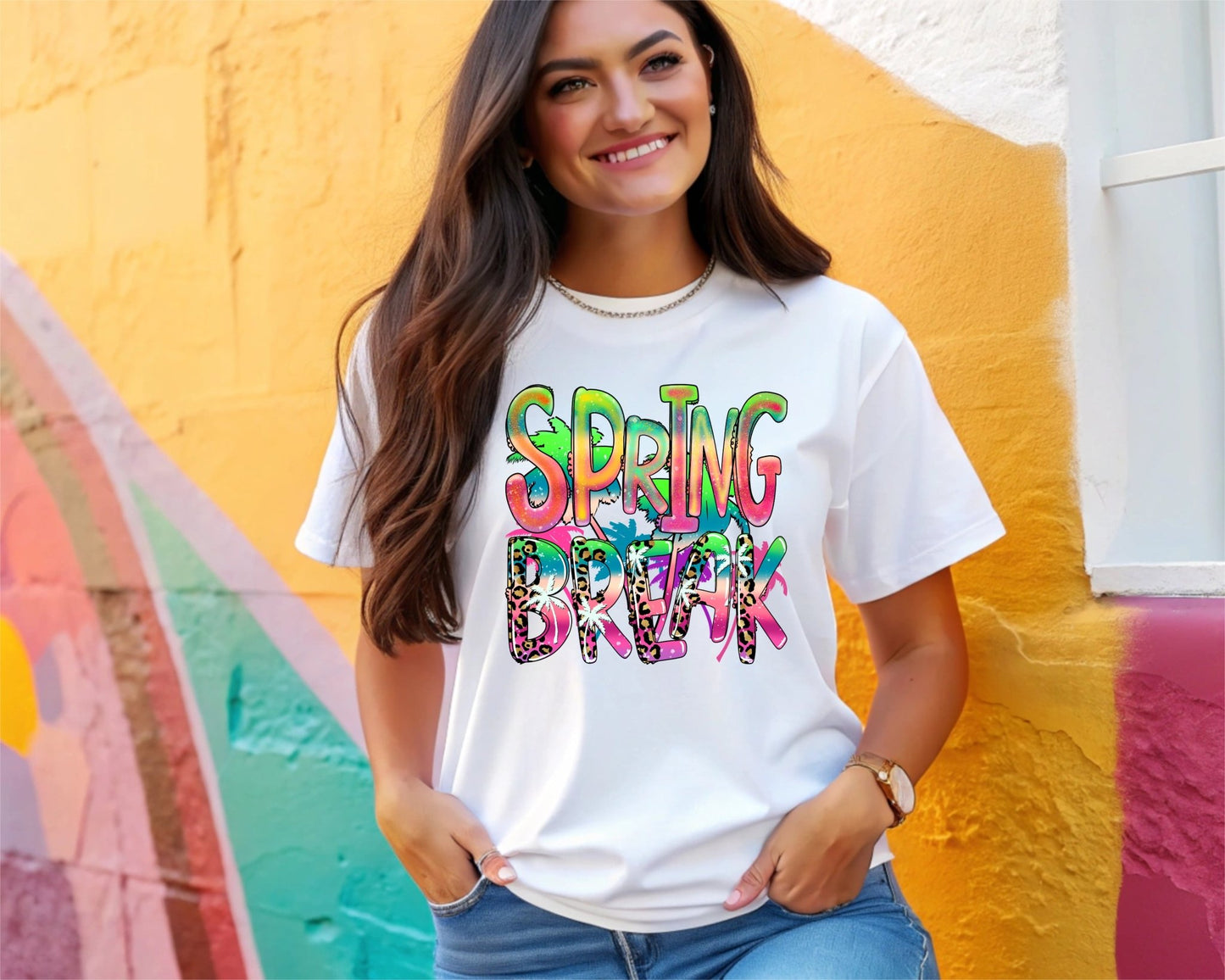 A smiling woman with long brown hair models the Spring Break t-shirt by DB Custom Creations, featuring colorful, patterned DTF-printed letters, while posing in front of a vibrant mural-painted wall.