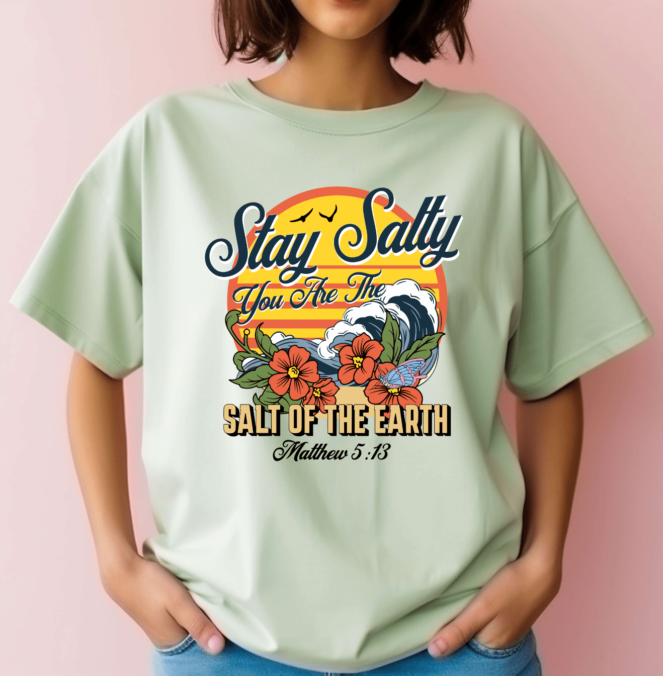 A woman wears the DB Custom Creations Stay Salty Bible verse light green T-shirt with colorful DTF waves, flowers, and “Stay Salty, You Are The Salt Of The Earth, Matthew 5:13” on a soft pink background.