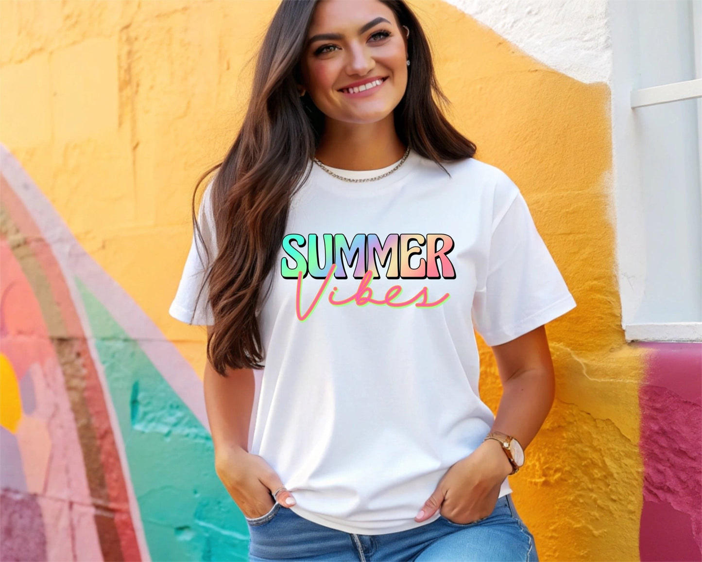 A smiling woman wears a white DB Custom Creations Summer Vibes t-shirt—featuring colorful letters printed with 11-inch DTF—standing by a vibrant, multicolored wall with her hands in her jeans pockets.