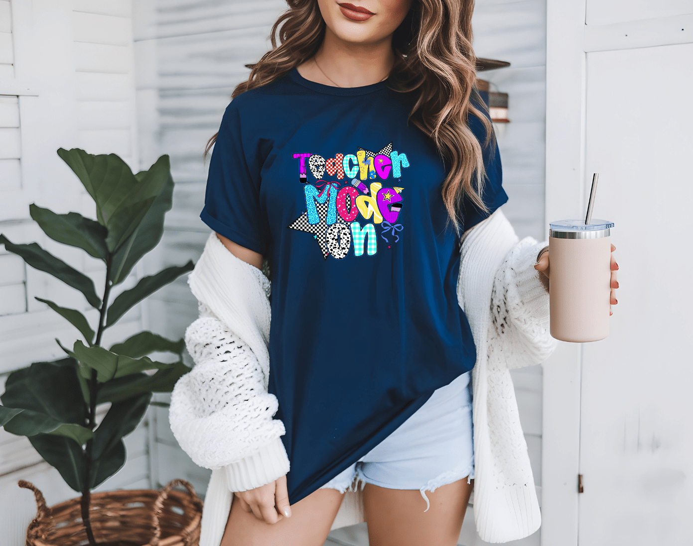 A woman wears the navy Teacher Mode graphic t-shirt by DB Custom Creations, paired with a white cardigan and denim shorts, holding a tumbler. She stands indoors near a plant, with white walls and shelves in the background.