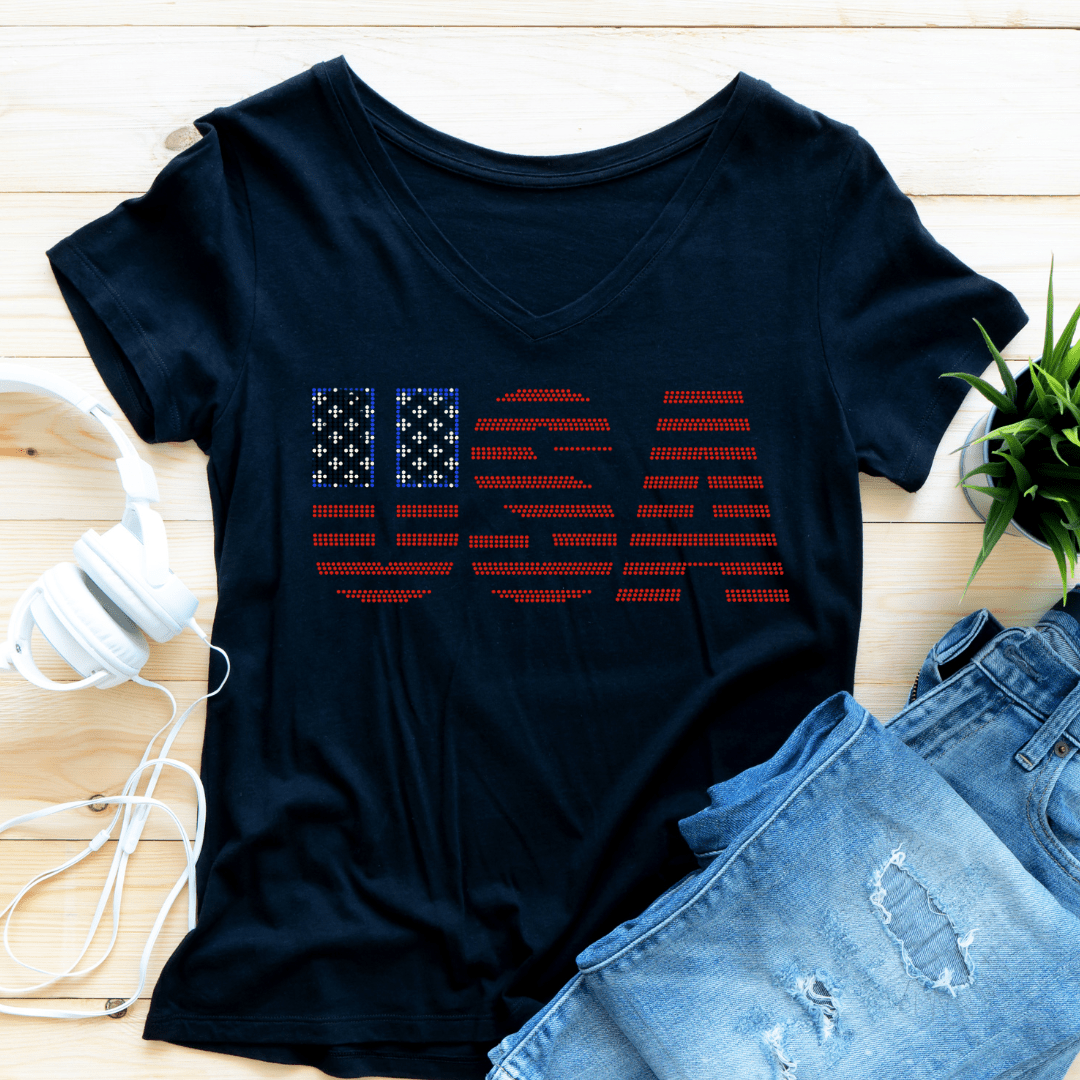 The DB Custom Creations Spangle Transfer - USA (9.5 x 4.5) decorates a black t-shirt with USA in red and white spangles; the U features flag-inspired stars and stripes, displayed with jeans, headphones, and a small green plant.