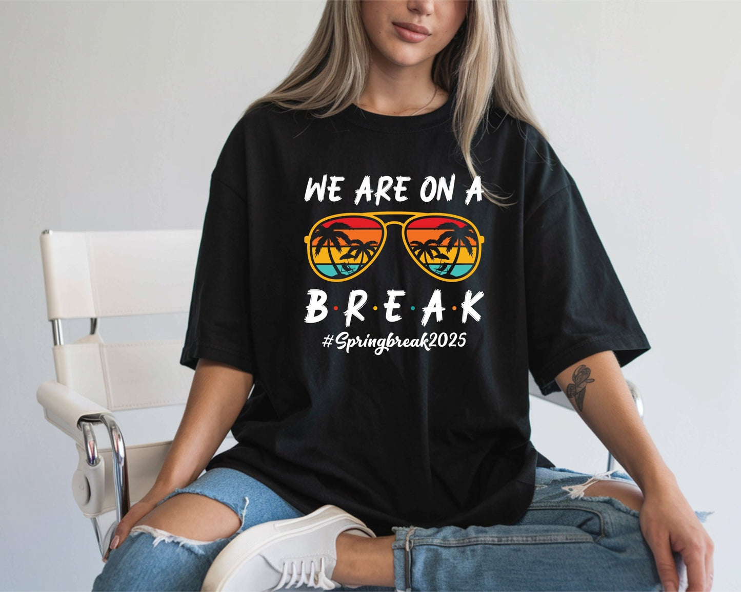 A woman in ripped jeans and a DB Custom Creations We Are On A Break 2025 black t-shirt, made with an 11 DTF heat transfer, wears sunset palm tree sunglasses while sitting on a white chair.