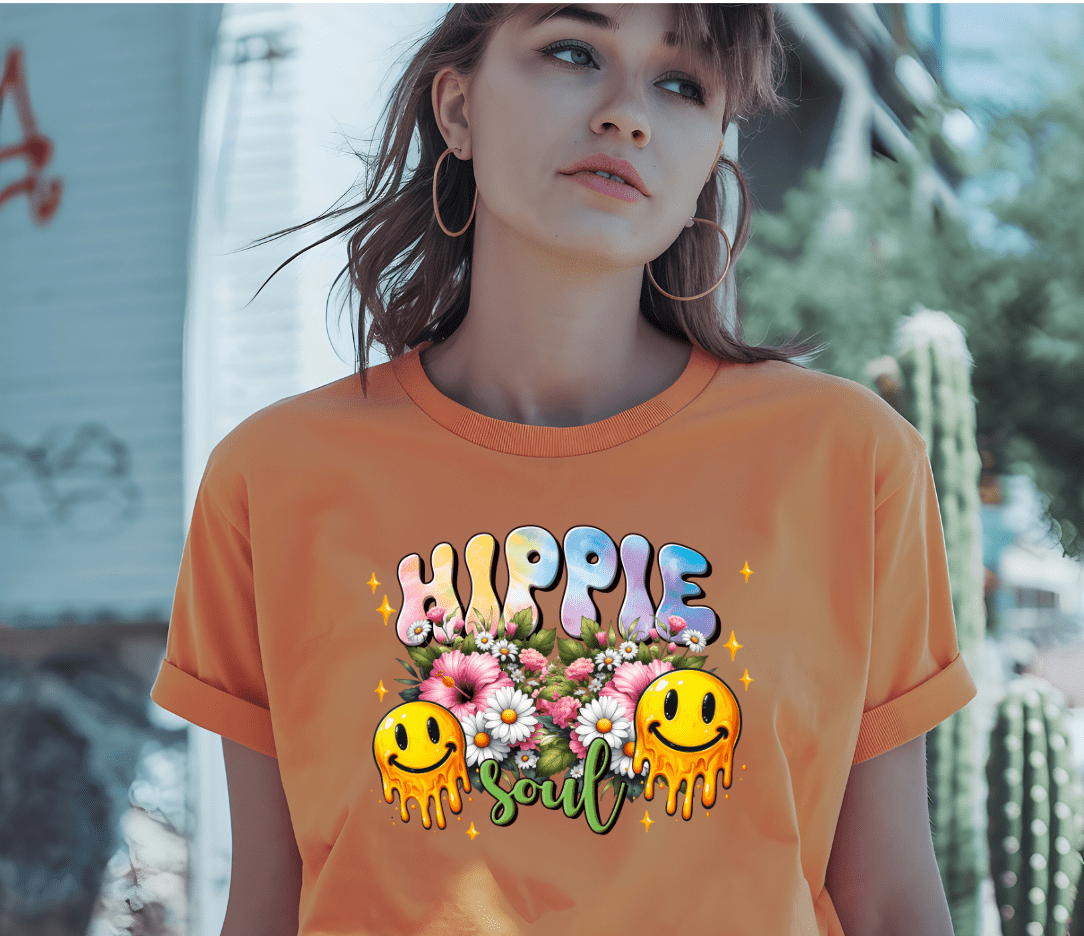 A young woman in hoop earrings wears the orange Hippie t-shirt by DB Custom Creations, featuring Hippie Soul, flowers, and melting smiley faces printed with vibrant DTF, as she stands outside by a cactus and graffiti wall.