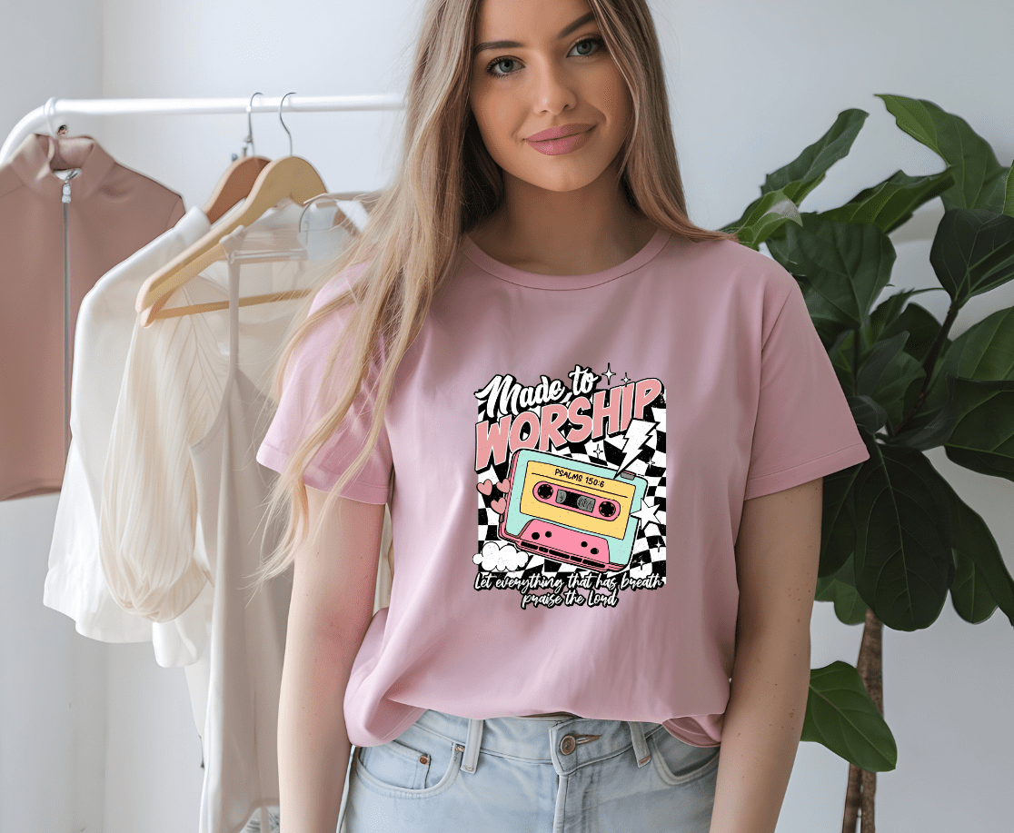 A woman with long blonde hair wears a light pink Made to Worship t-shirt by DB Custom Creations, featuring a colorful cassette tape graphic with a checkered background. She stands indoors near hanging clothes and a plant.