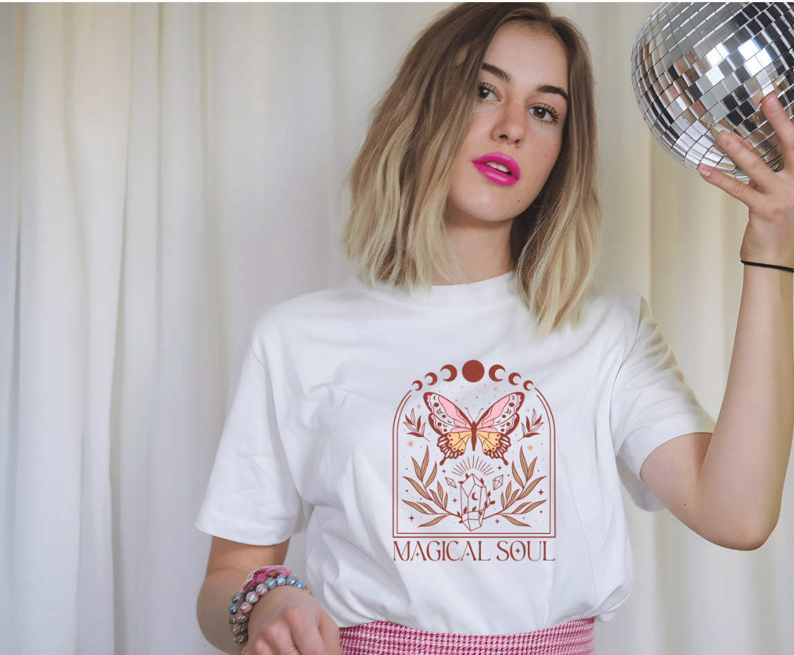 A young woman with a blonde bob and pink lipstick holds a disco ball, wearing the Magic Soul t-shirt by DB Custom Creations, featuring a mystical butterfly and flower design in 11-inch DTF print, plus pastel bead bracelets against white curtains.