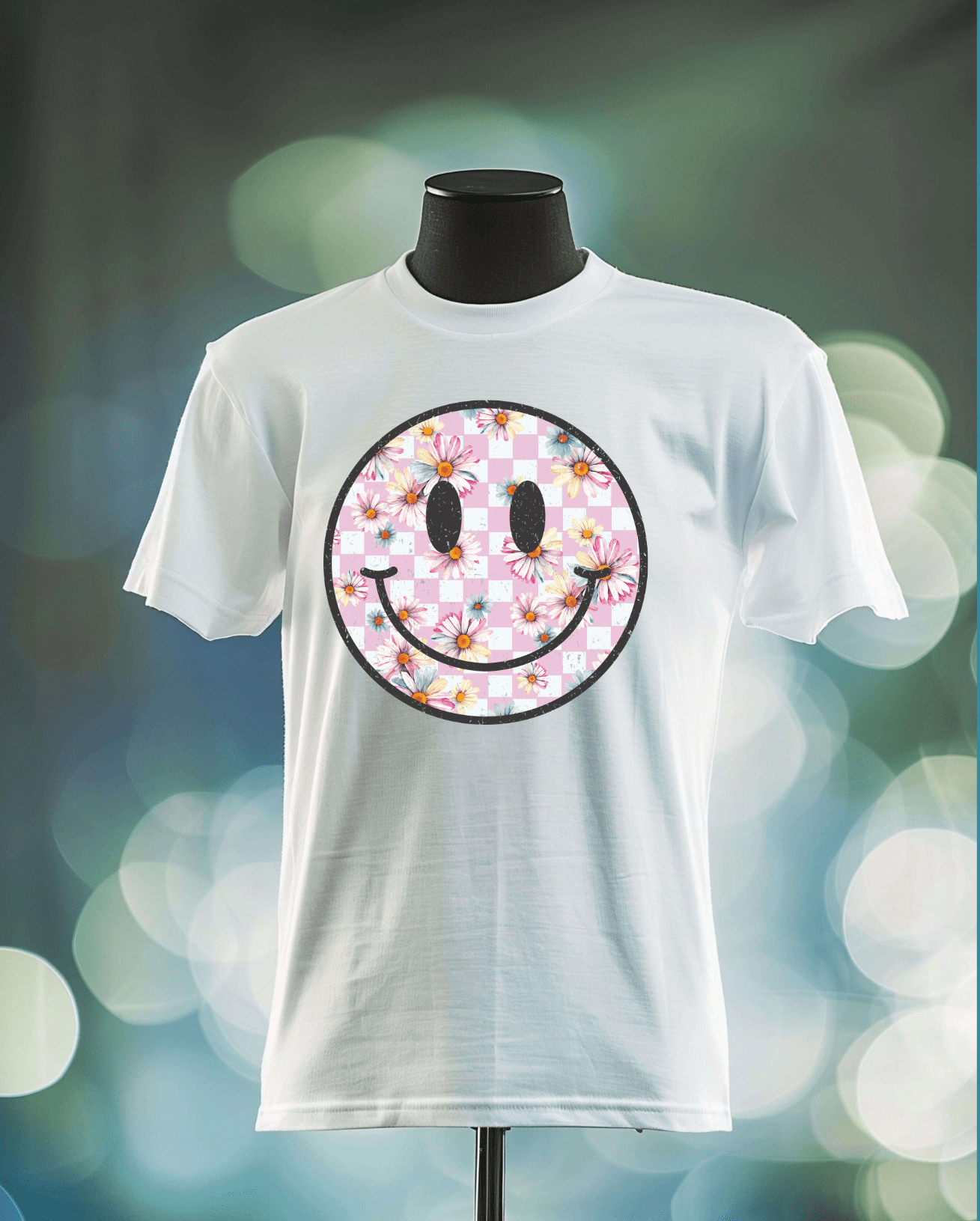 The Smiley Face t-shirt by DB Custom Creations features a large black-outlined smiley with a pink and white checkered fill and colorful floral accents, printed in vibrant DTF. Displayed on a mannequin against a blurred lights background.