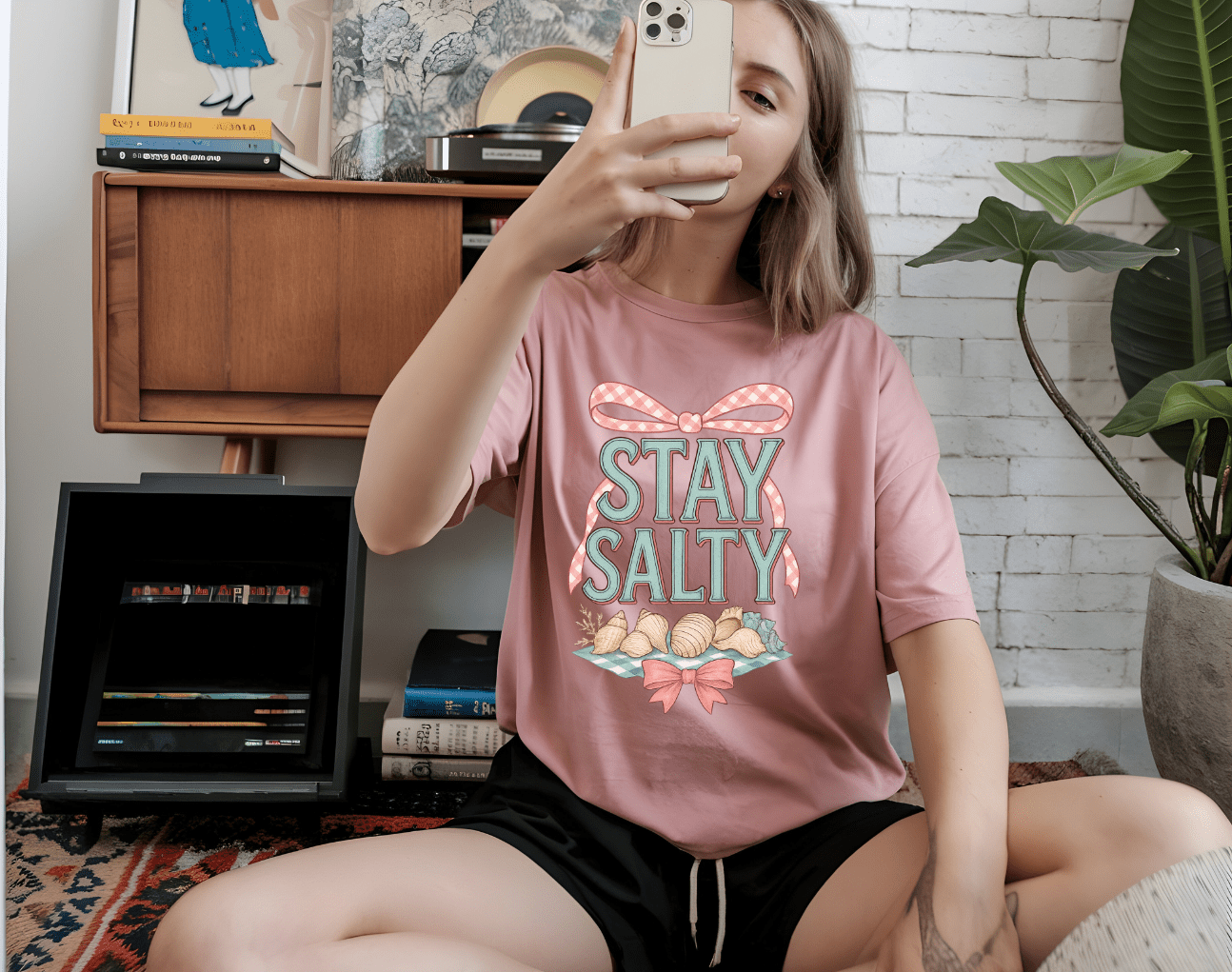 A woman sits on the floor taking a selfie, wearing DB Custom Creations stay salty pink t-shirt featuring a seashell and ribbon DTF Transfer design, in a cozy room with books, plants, and a record player.