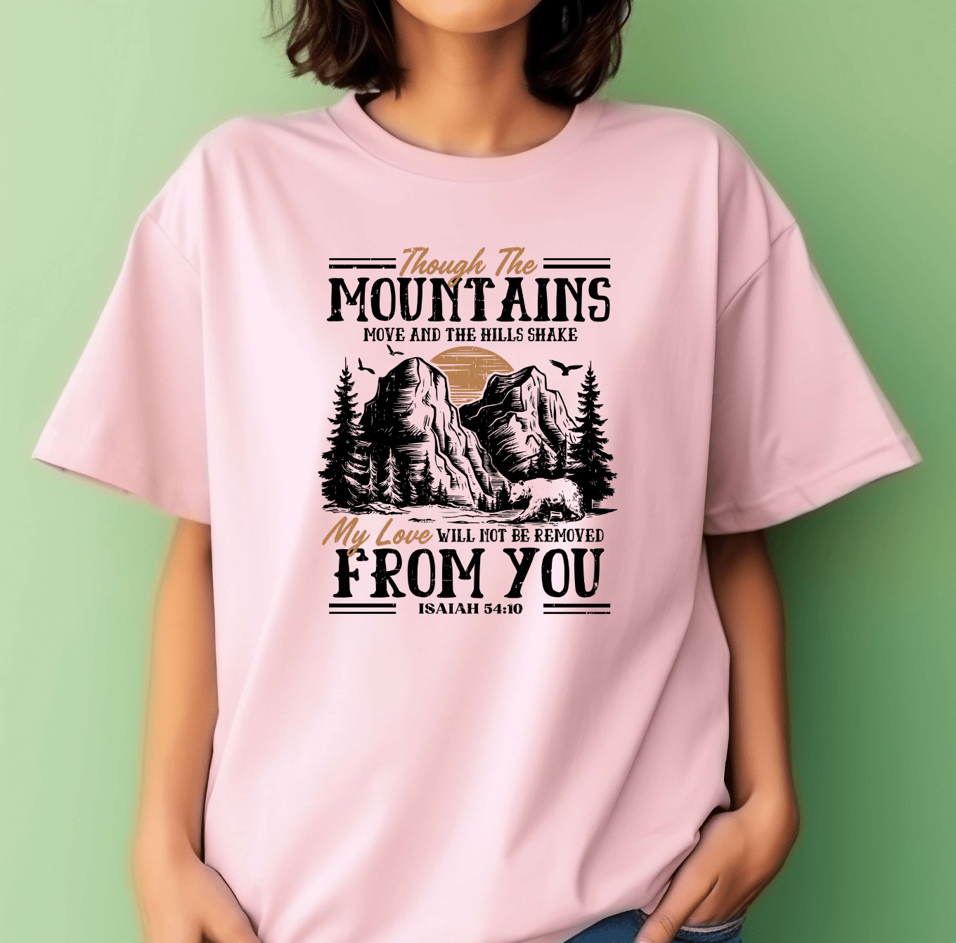 A woman wears the Through the Mountain pink t-shirt by DB Custom Creations, made with DTF printing. It features mountains and trees with the phrase: Though the mountains move, my love will not be removed from you. Isaiah 54:10.