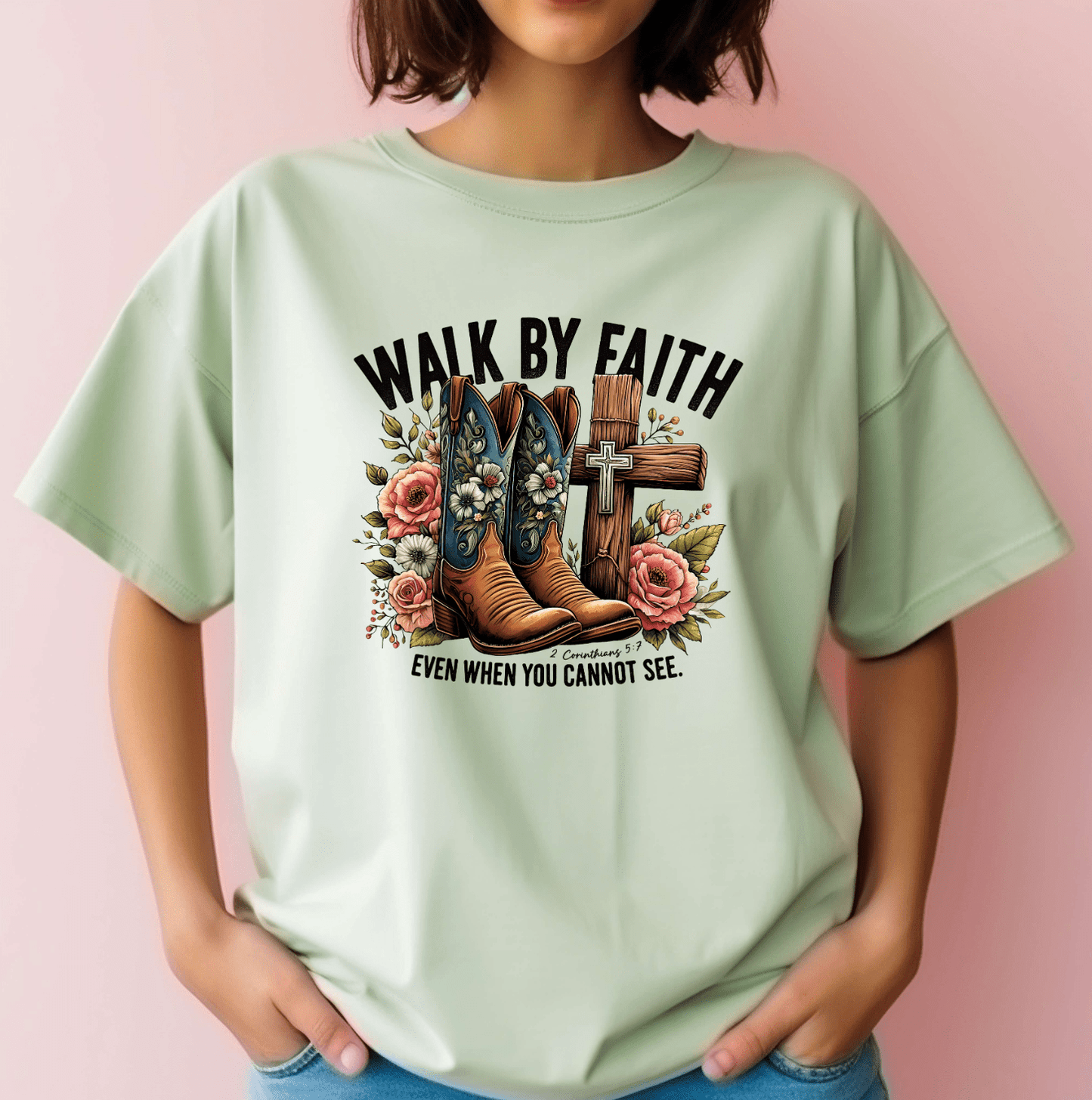 A person with short brown hair stands against a pink background, wearing the Walk by Faith light green tee from DB Custom Creations, featuring DTF-printed cowboy boots, flowers, a cross, and the phrase WALK BY FAITH EVEN WHEN YOU CANNOT SEE.