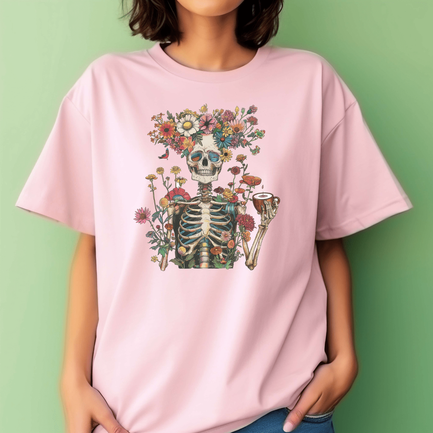 A person wears DB Custom Creations’ Whimsical Skeleton light pink tee, featuring a skeleton with a pipe and colorful flowers in 11-inch vibrant DTF print. The background is a plain light green wall.
