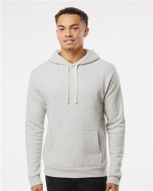 A young man wears the Next Level 9302, a light gray cotton-polyester fleece hoodie with a front pocket and drawstrings, standing against a plain white background.