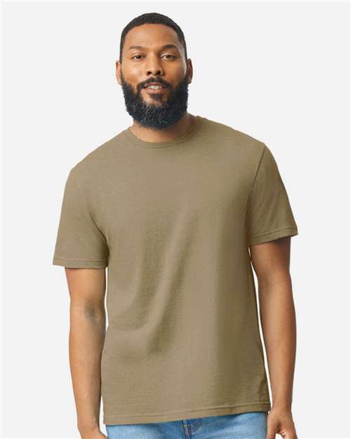 A bearded man with short hair wears a plain olive green Gildan Softstyle 64000CVC t-shirt and light blue jeans, standing against a neutral background. He faces forward with a relaxed expression. Brand: Gildan.