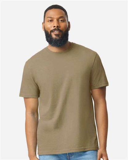 A bearded man with short hair wears a plain olive green Gildan Softstyle 64000CVC t-shirt and light blue jeans, standing against a neutral background. He faces forward with a relaxed expression. Brand: Gildan.
