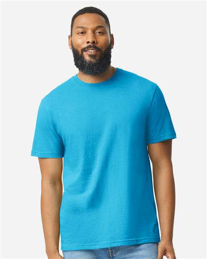 A bearded man with short hair wears a bright blue Gildan Softstyle 64000CVC t-shirt made from ring-spun cotton and light blue jeans, standing facing forward against a plain, light-colored background.