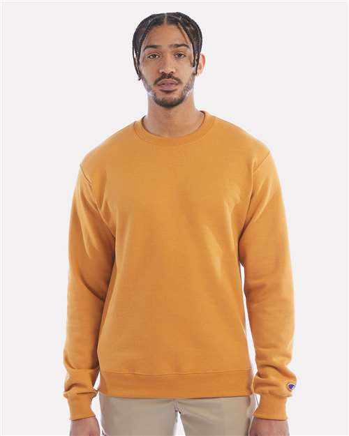 A man with braided hair stands facing forward, wearing a Champion S600 mustard yellow sustainable sweatshirt made from recycled polyester and beige pants against a plain light background.