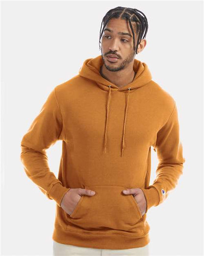 A man with braided hair wears the Champion S700 hoodie, made from recycled polyester, standing against a plain light background with hands in his pockets—showcasing sustainable style.