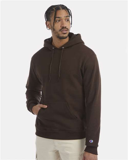 A man with braided hair wears the Champion S700 dark brown hoodie made from recycled polyester, paired with light pants. He stands against a plain light background, one hand in his pocket, looking slightly to the side.