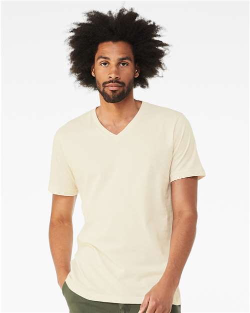 A man with medium brown skin and curly afro hair wears the BELLA + CANVAS 3005 unisex V-neck in light beige, made from Airlume combed cotton, and dark green pants. He stands against a white background with a neutral expression.